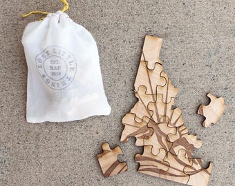 May include: A wooden jigsaw puzzle shaped like the state of Idaho with a sunburst design. The puzzle is in a white drawstring bag with the text "Lost Little Things Est. Mar 2010" printed on it.