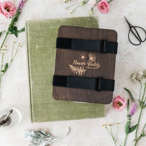 May include: A wooden flower press with black straps, the text "Flower Buddy Made in USA", and a green book. Pink flowers and small scissors are arranged around the press. The press is designed for preserving flowers.
