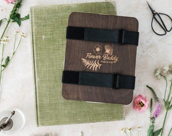 May include: A wooden flower press with black straps, the text "Flower Buddy Made in USA", and a green book. Pink flowers and small scissors are arranged around the press. The press is designed for preserving flowers.