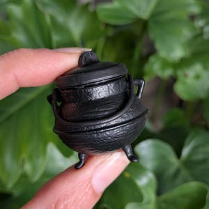 May include: A small, black, round cauldron with a textured surface, three short legs, a matching lid with a central handle, and a curved handle attached to the sides. The exterior features embossed markings around the upper rim area.