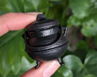 May include: A small, black, round cauldron with a textured surface, three short legs, a matching lid with a central handle, and a curved handle attached to the sides. The exterior features embossed markings around the upper rim area.