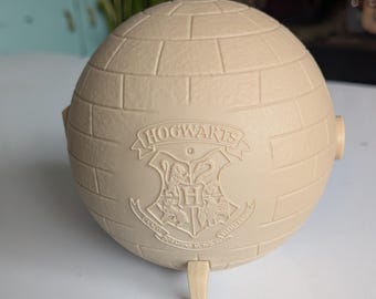 May include: A spherical, beige-colored object with a textured brick pattern across its surface. The front features an embossed Hogwarts crest with the text HOGWARTS above it and the Latin motto Draco Dormiens Nunquam Titillandus below it.
