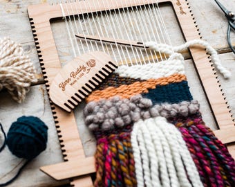 May include: A wooden loom with a colorful woven tapestry hanging from it. The loom is labeled "Yarn Buddy Made in USA". There is a wooden comb with the same label next to the loom.