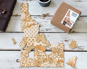 May include: A wooden jigsaw puzzle of the state of Idaho, with each piece featuring a different pattern. The puzzle is assembled on a white wooden surface, with a brown box labeled "Idaho Puzzle" in the top right corner.