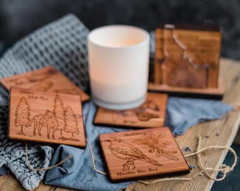 May include: Set of four wooden coasters with laser-engraved images of animals and plants. The coasters feature a white-tailed deer, an Appalachian tree, a mountain bluebird, and a rhododendron. The coasters are made of natural wood and have a rustic look.