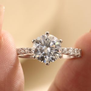 May include: A silver-toned ring featuring a large, round, brilliant-cut clear center stone held by six prongs. The band is accented with a row of smaller, round, clear pavé-set stones along each side, creating a sparkling, reflective appearance.