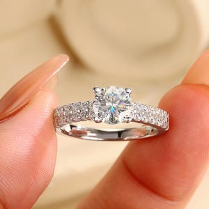 May include: A silver-toned ring featuring a prominent round-cut center stone held by four prongs. The band is crafted with a pavé setting, displaying multiple small, sparkling stones embedded along the surface of the metal shank on both sides.