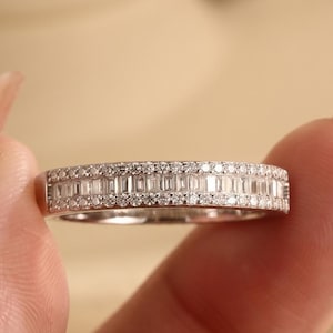 May include: A silver-toned band ring featuring a central row of baguette-cut stones flanked by two outer rows of small, round, brilliant-cut stones. The metal band has a polished finish and a uniform width, showcasing a multi-faceted, sparkling design.