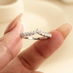 May include: A silver-toned V-shaped chevron ring featuring a row of small, clear, round-cut stones set along the top edge. The band is thin and polished, showcasing a symmetrical design that comes to a point at the center of the ring.