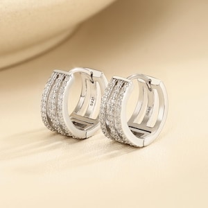 May include: A pair of silver-toned hoop earrings featuring three parallel rows of small, clear, sparkling stones. Each hoop has a hinged closure and is stamped with the text S925 on the inner side, indicating the metal composition of the jewelry.