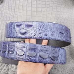 May include: A blue belt featuring a textured crocodile skin pattern with raised scales. The belt displays a distinct, organic hide texture with varying scale sizes across its surface, showcasing a deep blue color with darker highlights.