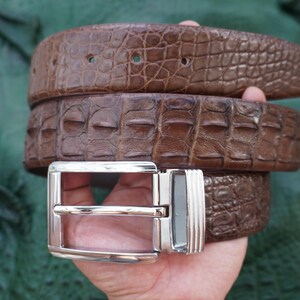 Peut inclure: Une ceinture en cuir marron avec une texture de peau de crocodile. La ceinture est enroul&eacute;e, montrant le motif et la texture d&eacute;taill&eacute;s. Elle a une boucle de couleur argent.