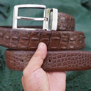 Peut inclure: Une ceinture en cuir marron avec une boucle argent&eacute;e. La ceinture pr&eacute;sente un motif textur&eacute; en forme de crocodile. La boucle est rectangulaire et a une finition brillante. La ceinture est tenue par une main.