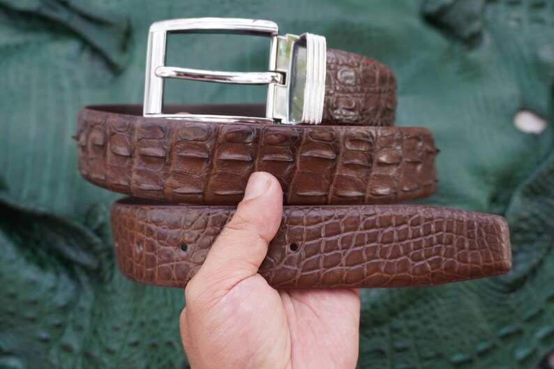 Peut inclure: Une ceinture en cuir marron avec une boucle argent&eacute;e. La ceinture pr&eacute;sente un motif textur&eacute; en forme de crocodile. La boucle est rectangulaire et a une finition brillante. La ceinture est tenue par une main.
