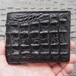 Puede incluir: Una cartera de cuero negro con un patrón de cocodrilo texturizado. La cartera es rectangular y parece estar sujeta en la mano de una persona. El cuero tiene un acabado brillante y la cartera es de un negro oscuro.