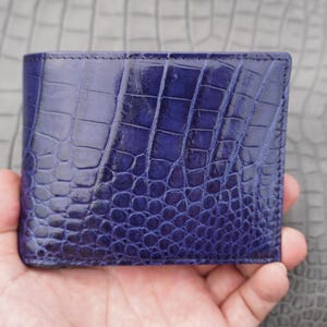 Peut inclure: Un portefeuille pliable d'un bleu profond avec un motif texturé en relief imitant l'apparence d'une peau de reptile exotique. Le portefeuille présente une forme rectangulaire avec des coutures visibles sur les bords et une finition brillante.