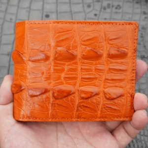 May include: A vibrant orange bifold wallet featuring a prominent, raised, and textured scale pattern across the exterior surface. The wallet has a rectangular shape with visible stitching along the edges and a glossy, smooth finish on the scales.