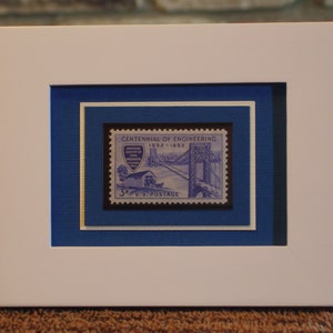 May include: A framed 3¢ U.S. postage stamp commemorating the Centennial of Engineering, 1852-1952. The stamp features a blue and white illustration of a bridge with a car crossing it. The stamp is surrounded by a white mat and a blue mat.