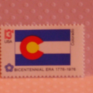 May include: A rectangular postage stamp depicting the Colorado state flag. The flag design includes a blue field, a white stripe, and a red 'C' with a yellow circle. The stamp is marked '13¢ USA' and 'Bicentennial Era 1776-1976'.