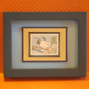 Puede incluir: Una imagen enmarcada de un perro de dibujos animados que lleva una camisa azul y sostiene una carta. La imagen está rodeada por una moldura azul y una moldura dorada. La imagen está en un marco azul oscuro.