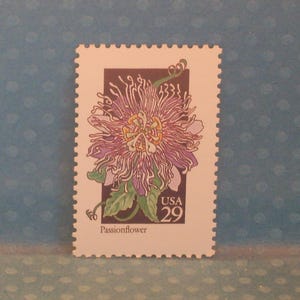 Puede incluir: Un sello postal con una ilustración de una flor de la pasión. El sello tiene un borde blanco dentado. La flor es morada, amarilla y verde, con el texto "USA 29" y "Passionflower".