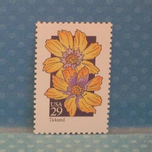Puede incluir: Un sello postal de EE. UU. con dos flores de Tickseed amarillas y naranjas con centros morados. El sello tiene un borde blanco con un borde dentado y el texto "USA 29 Tickseed".