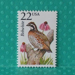 May include: A 22 cent USA postage stamp featuring a Bobwhite Quail with brown and white feathers, perched on a branch with pink flowers. The stamp has a white background with a green border.