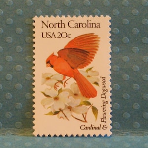 May include: A vintage postage stamp from North Carolina, USA, featuring a vibrant red cardinal bird with outstretched wings perched on white flowering dogwood blossoms. The stamp is valued at 20 cents.