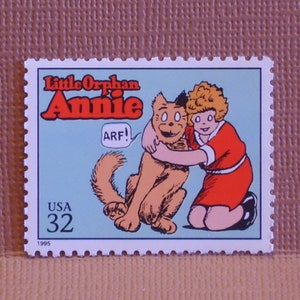May include: A vintage US postage stamp featuring a cartoon image of Little Orphan Annie with her dog Sandy. The stamp is blue and white with the text "Little Orphan Annie" and "USA 32" printed on it. The year 1995 is printed in the bottom left corner.
