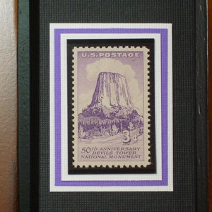 Könnte beinhalten: Eine gerahmte US-Briefmarke aus der Zeit mit dem Devils Tower National Monument in Wyoming. Die Marke ist lila und zeigt ein Bild des Turms mit Bäumen im Vordergrund. Die Marke ist von einem weißen und einem lilafarbenen Rahmen umgeben. Der Text auf der Marke lautet "U.S. POSTAGE 3¢ 50th Anniversary Devils Tower National Monument".