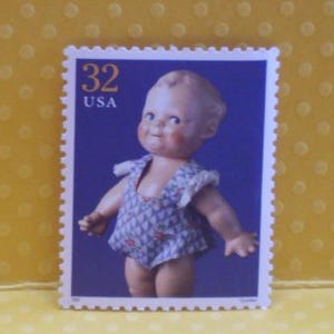 Op de afbeelding: Een vintage postzegel met een pop in een blauw en wit patroon. De postzegel heeft een blauwe achtergrond en de tekst "32 USA". De pop heeft roze wangen en staat.