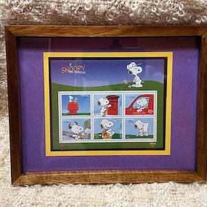 Puede incluir: Una impresión enmarcada de una colección de sellos de Peanuts con Snoopy en varias poses. Los sellos están rodeados por un borde amarillo y una montura morada. El marco es de madera oscura.