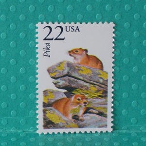 May include: A postage stamp featuring two brown pikas on a rocky landscape. The stamp has the text "22 USA" at the top and "Pika" on the left side. The background is a teal color with a dotted pattern.
