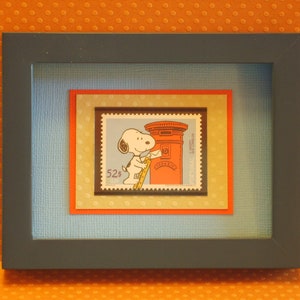 Puede incluir: Obra de arte enmarcada con un sello postal con Snoopy, un perro blanco, apoyado en un buzón rojo. El sello está sobre un fondo azul, con un borde rojo y amarillo, y un marco gris oscuro.