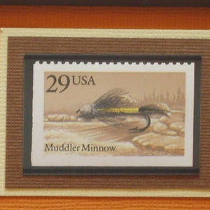 Pode incluir: Um selo postal vintage com uma mosca marrom, preta e amarela em um fundo branco. O selo é rotulado como "29 USA" e "Muddler Minnow".