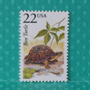 May include: A postage stamp featuring a detailed illustration of a box turtle. The stamp reads "22 USA" and "Box Turtle". The turtle has a brown and orange shell, with green foliage in the background.