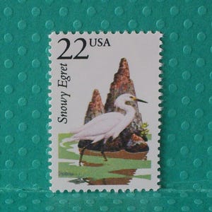 May include: A postage stamp featuring a Snowy Egret, a white bird with a black beak and legs, standing in a marshy area. The stamp has a value of 22 cents and the text "USA" and "Snowy Egret". The background includes brown rock formations.