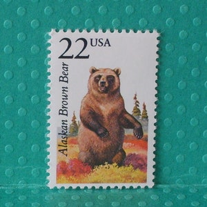 May include: A 22 cent USA postage stamp featuring a brown Alaskan brown bear standing in a field of wildflowers. The stamp has a white background with a blue border.