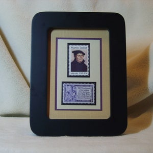 May include: A framed display of two vintage postage stamps featuring Martin Luther. The top stamp shows a portrait of Martin Luther in a black robe and a white hat. The bottom stamp shows a scene of the Reformation with a church in the background. Both stamps are purple and have the text "USA 20c" printed on them.