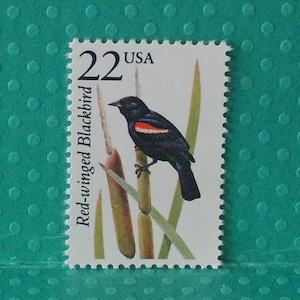May include: A postage stamp featuring a Red-winged Blackbird perched on reeds. The bird is black with a red and yellow shoulder patch. The stamp has the text "22 USA" and "Red-winged Blackbird".
