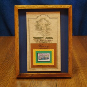 May include: A framed print of a vintage postcard featuring a geyser erupting in Yellowstone National Park, Wyoming. The print includes a small, framed stamp with a picture of the park.