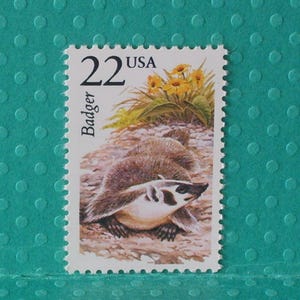 May include: A vintage postage stamp featuring a badger illustration. The stamp has a white border with the text "22 USA" and "Badger". The badger is depicted in shades of brown, white, and black, with yellow flowers in the background.