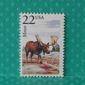 May include: A postage stamp featuring a brown moose with large antlers walking across a landscape of rocks and colorful vegetation. The stamp reads "22 USA" and "Moose" in black text.