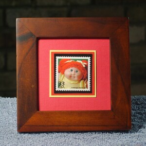 May include: A framed Cabbage Patch Kid postage stamp with a red background and a black and yellow border. The stamp features a Cabbage Patch Kid doll with red hair and a yellow dress. The stamp is from 1993 and says "USA" and "Cabbage Patch Kids" on it.
