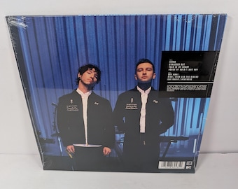 May include: A sealed vinyl record album featuring a blue-toned photograph of two individuals in black jackets standing before a vertical-slatted background. A black rectangular tracklist sticker is affixed to the upper right corner of the plastic wrap.