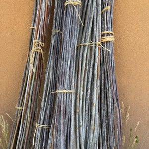 May include: A bundle of slender, gray and brown wooden sticks tied together with twine. The sticks are arranged vertically against a textured, tan-colored wall. Some dried grass is visible at the bottom.