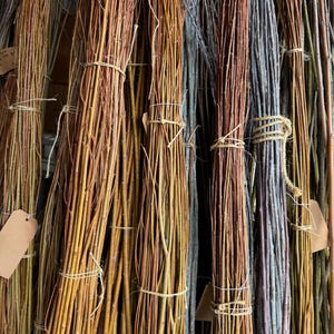 May include: Bundles of willow branches in various colors, including yellow, brown, and gray. The branches are tied together with twine and have small, blank tags attached. The natural materials are arranged closely together.