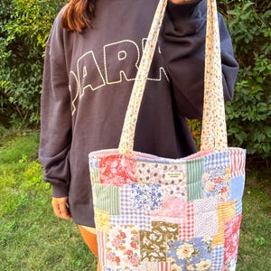 May include: A quilted patchwork tote bag featuring a variety of floral, gingham, and striped fabric squares in shades of pink, blue, green, and yellow. The bag has two long shoulder straps and a small rectangular label reading Spruce House Studio.