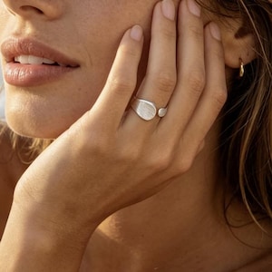 Könnte beinhalten: Ein silberner Ring mit einer glatten, unregelmäßigen Form. Der Ring hat eine große, oval geformte Oberseite und einen kleineren, runden Akzent. Der Ring wird an einem Finger getragen, mit dem Meer im Hintergrund.