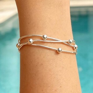 Könnte beinhalten: Ein silbernes Armband mit drei Strängen, die jeweils mit kleinen, runden Kugeln verziert sind. Das Armband wird an einem Handgelenk getragen, mit einem verschwommenen Hintergrund aus Pool und Außenbereich. Das Design des Armbands ist schlicht und elegant.